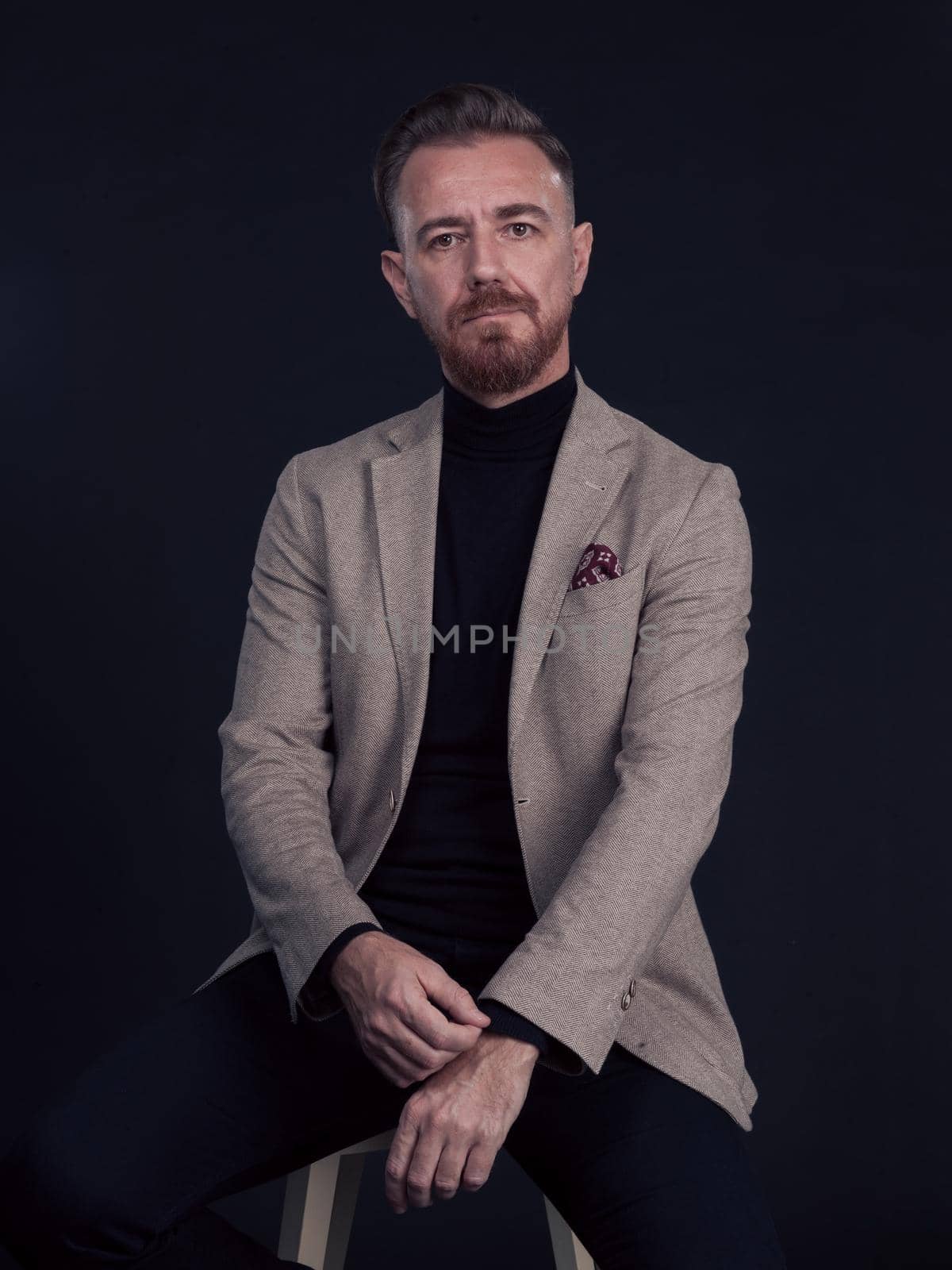 Portrait of adult businessman wearing trendy suit and sitting in modern studio on stylish chair against the black background. Horizontal mockup. High quality photo