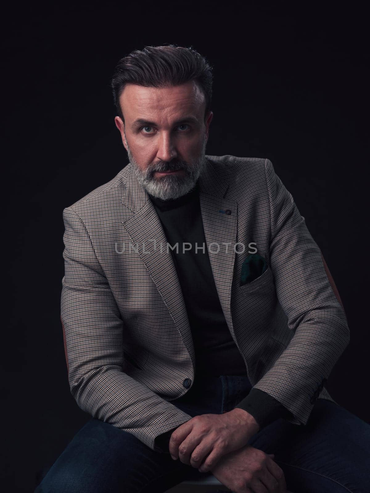 Portrait of adult businessman wearing trendy suit and sitting in modern studio on stylish chair against the black background. Horizontal mockup. High quality photo