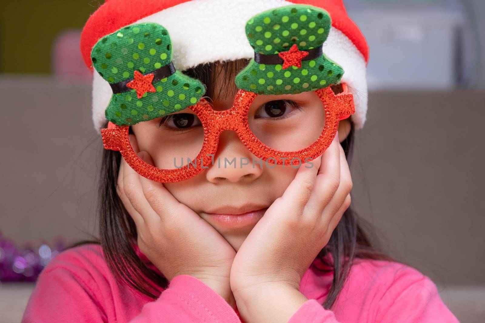 Little girl in a Santa hat sitting on the floor and making wishes near the Christmas tree at home. Happy New Year and Merry Christmas.