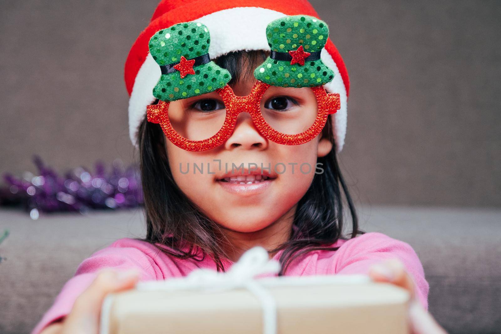 Happy little girl in Santa hat giving a Christmas present at home. Happy New Year and Merry Christmas.