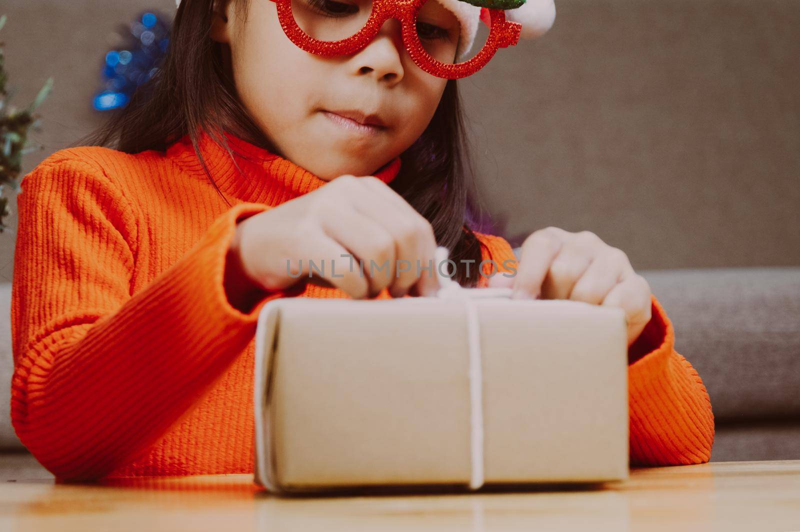 Little girl in Santa hat opening gift box sitting in the living room with Christmas tree at home. Happy New Year and Merry Christmas.