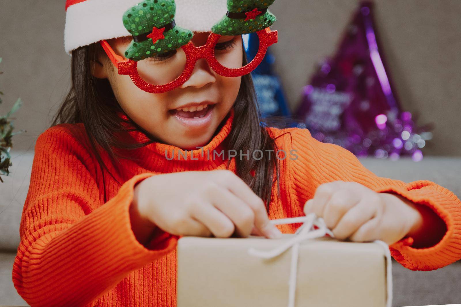 Little girl in Santa hat opening gift box sitting in the living room with Christmas tree at home. Happy New Year and Merry Christmas.