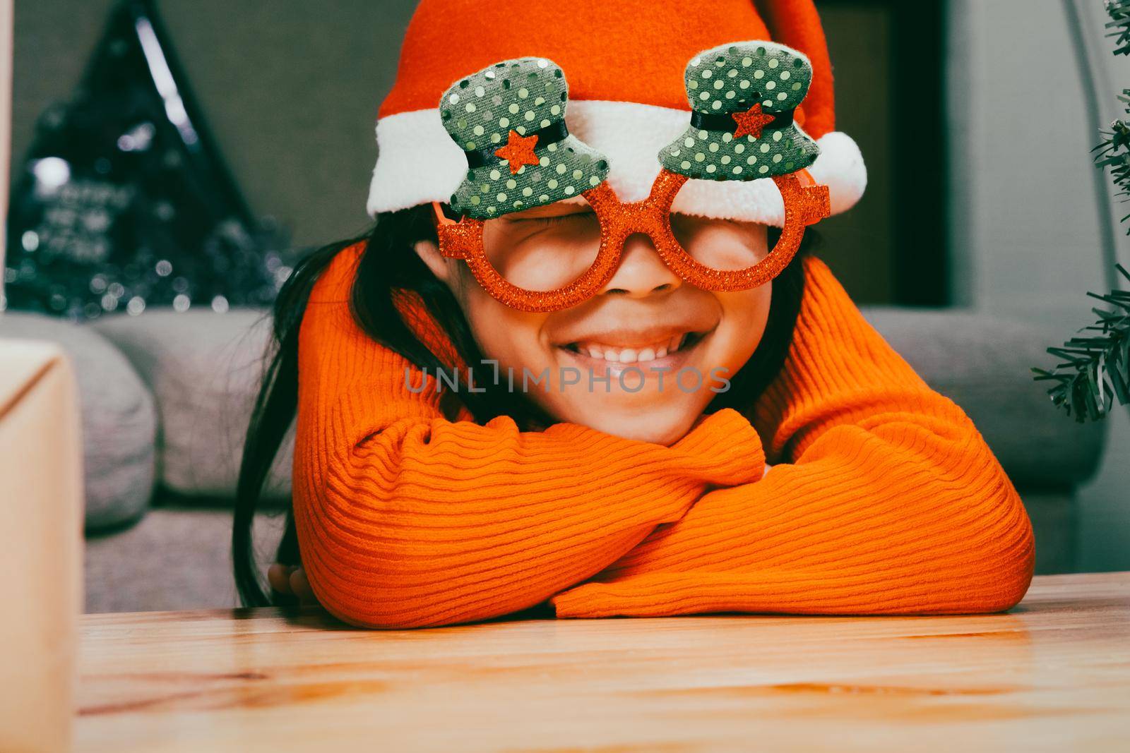 Little girl in a Santa hat sits with her eyes closed and making wishes near the Christmas tree indoors. Happy New Year and Merry Christmas.
