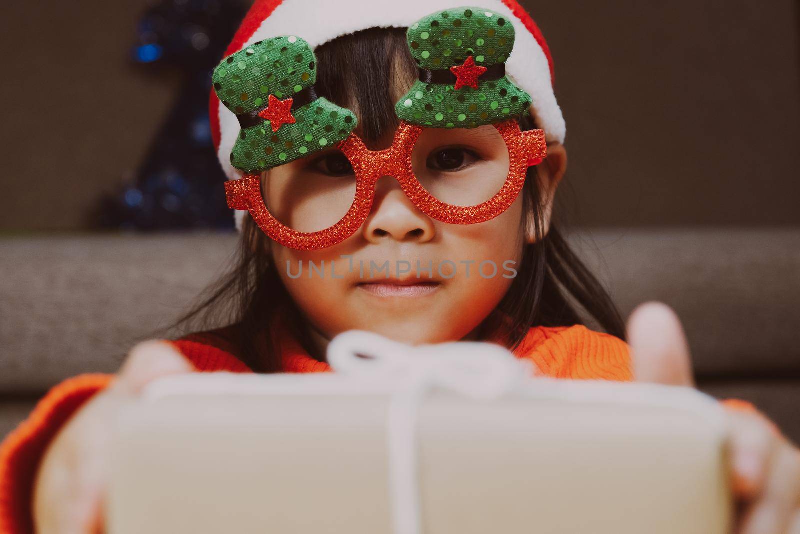 Happy little girl in Santa hat giving a Christmas present at home. Happy New Year and Merry Christmas.