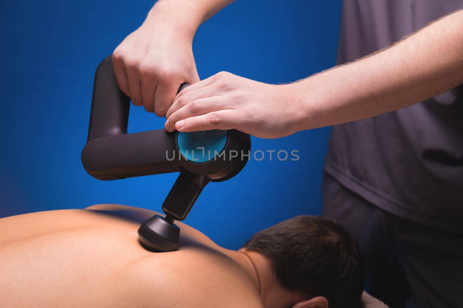 Close-up of a professional male masseur stimulates the back muscles of a male patient in a dark spa room for massage. Percussion mechanical effect on overstrained muscles by yanik88