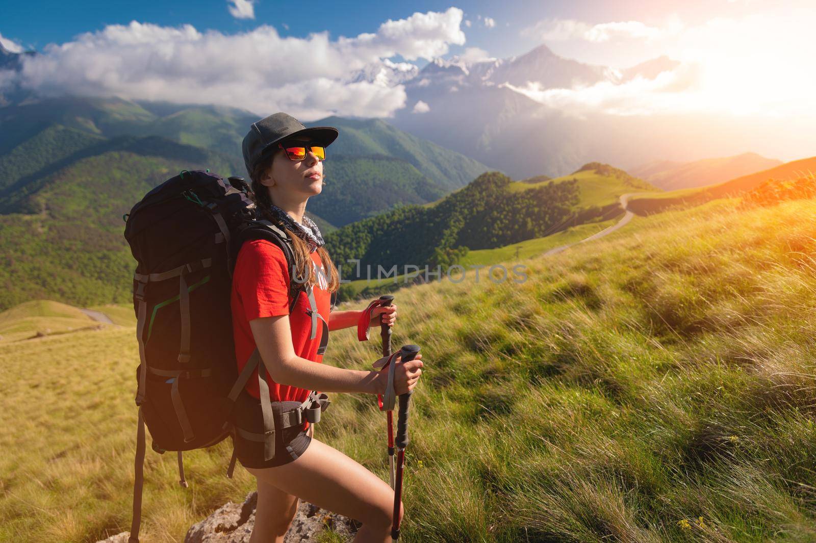 A tourist girl with a backpack admires the sunset from the mountainside. Traveler in the background of mountains and clouds at sunset. Woman tourist in sunglasses, cap and with a backpack by yanik88