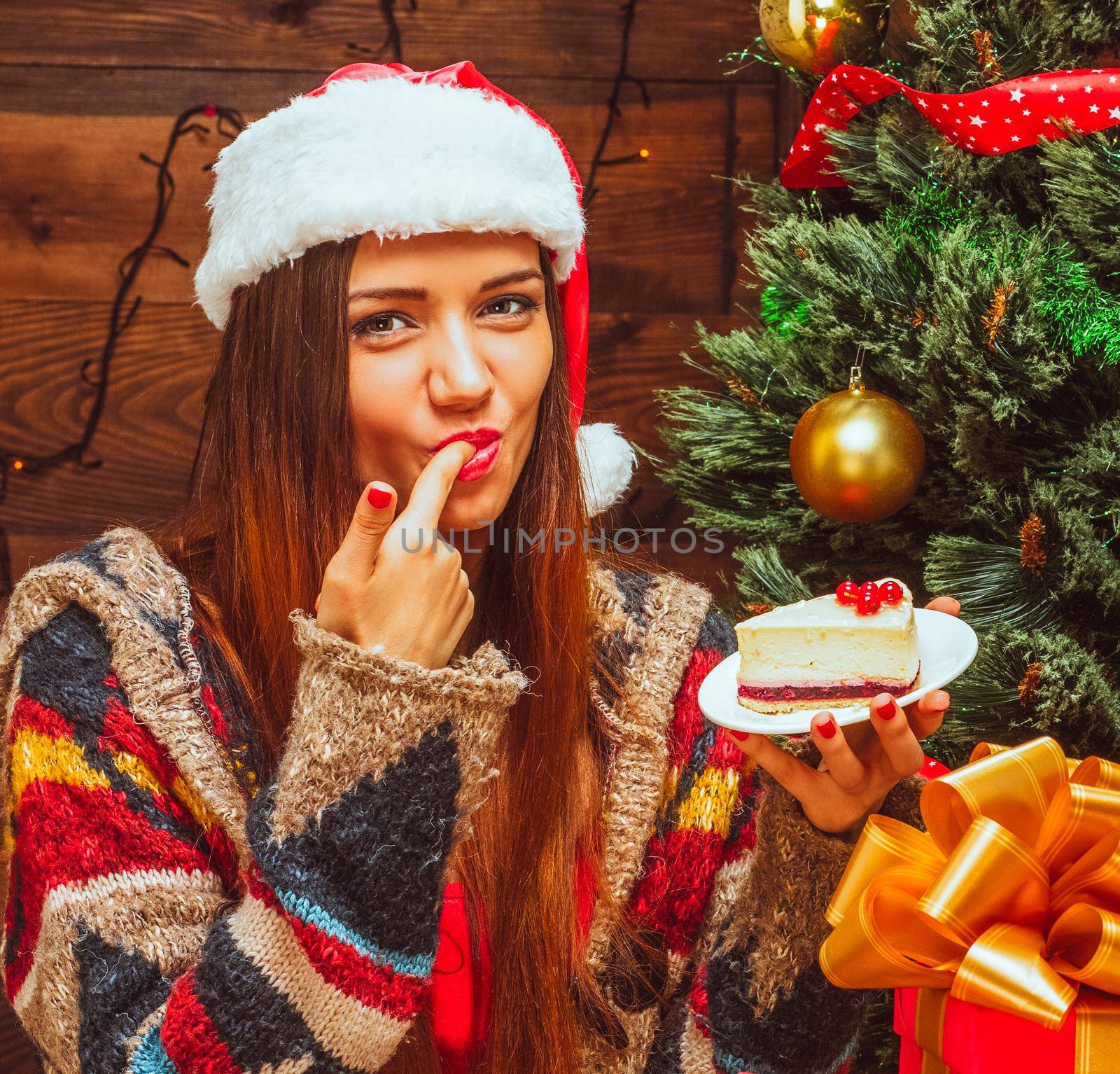 Bright lady holding a plate with a slice of cake and tasting it, finger in the mouth, Christmas Eve, dinner, cake, Tree, Christmas Gifts, Wood Background. Close-up. High quality photo
