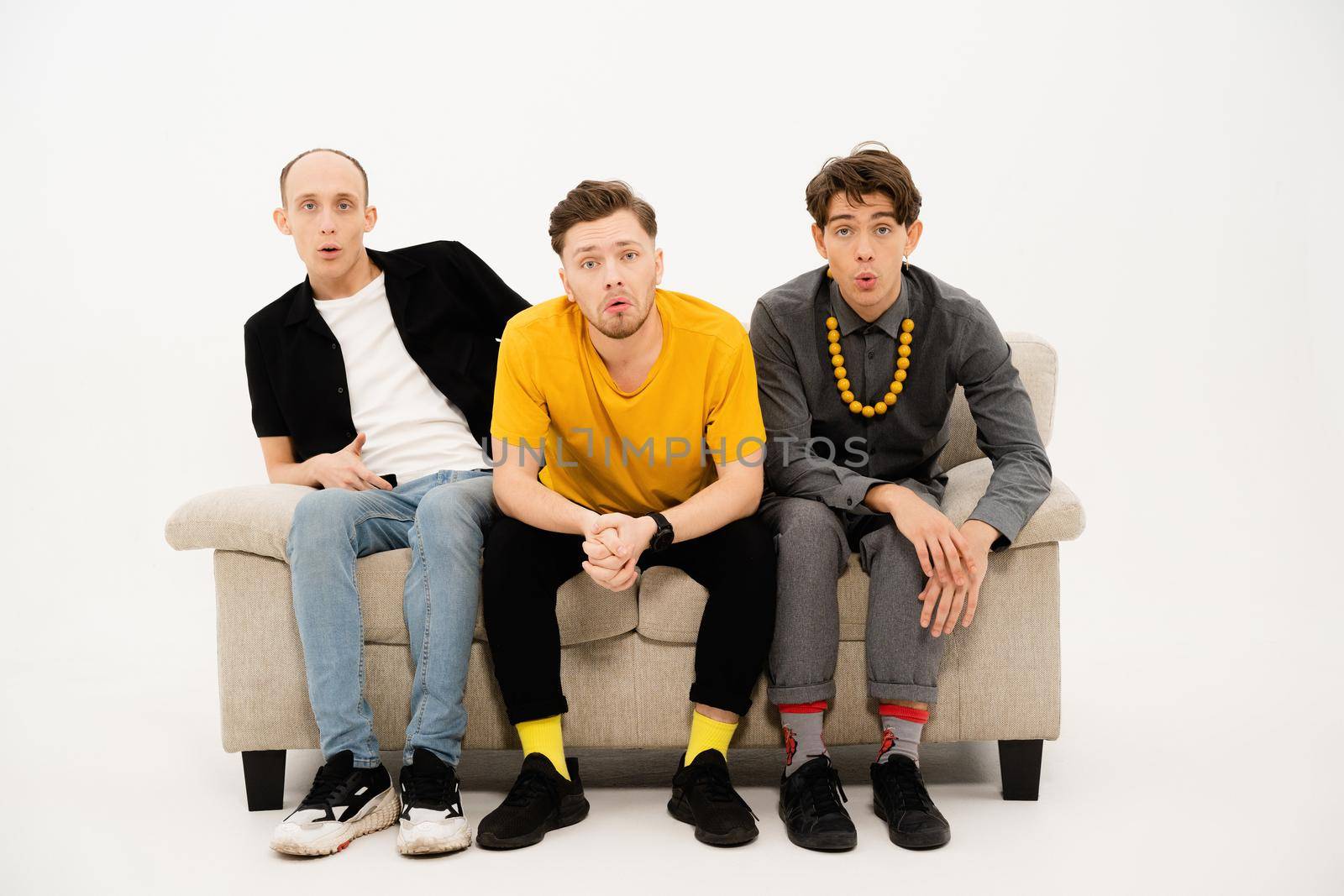 Portrait of three young friends spend time together sitting on the small sofa looking at the camera isolated on white background. Group of guys sitting on couch and communicates on white.