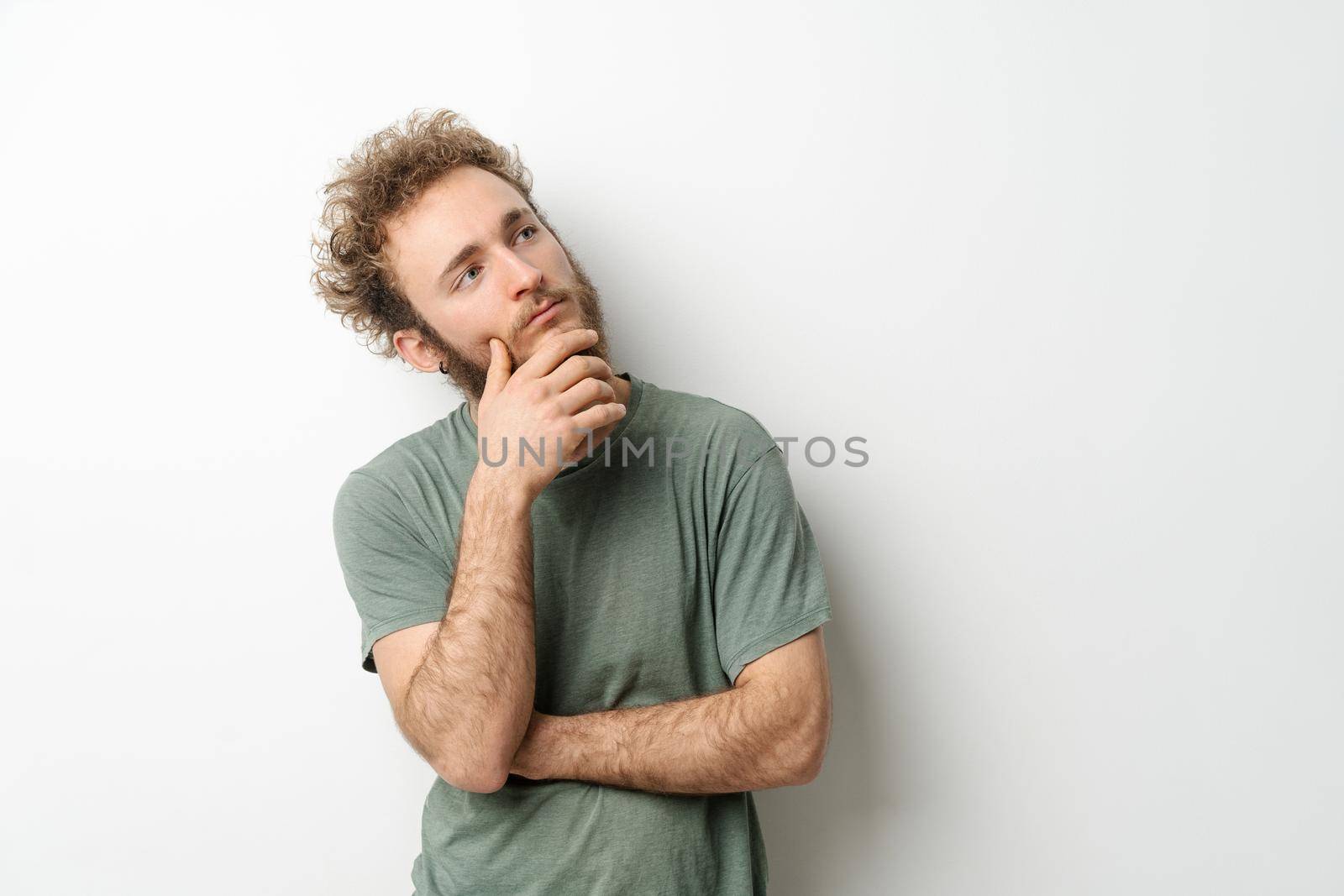 Pensive look of a handsome young man with curly hair in olive t-shirt looking upwards isolated on white background. Portrait of smiling young man with hands folded.