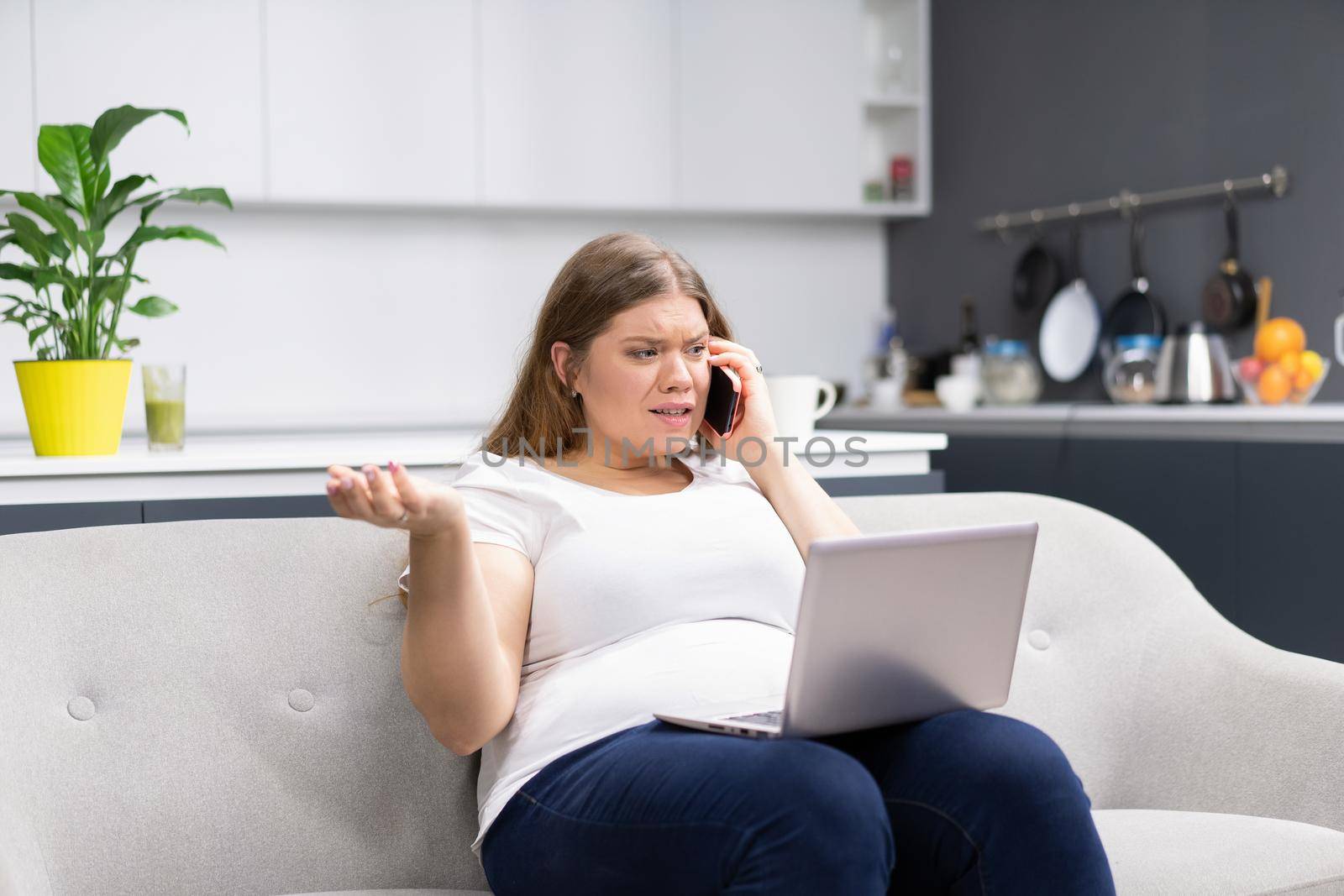 Talking on the phone while working on laptop sitting at home overweight young woman staying at home during quarantine. Self isolation. Working distantly from home using laptop.