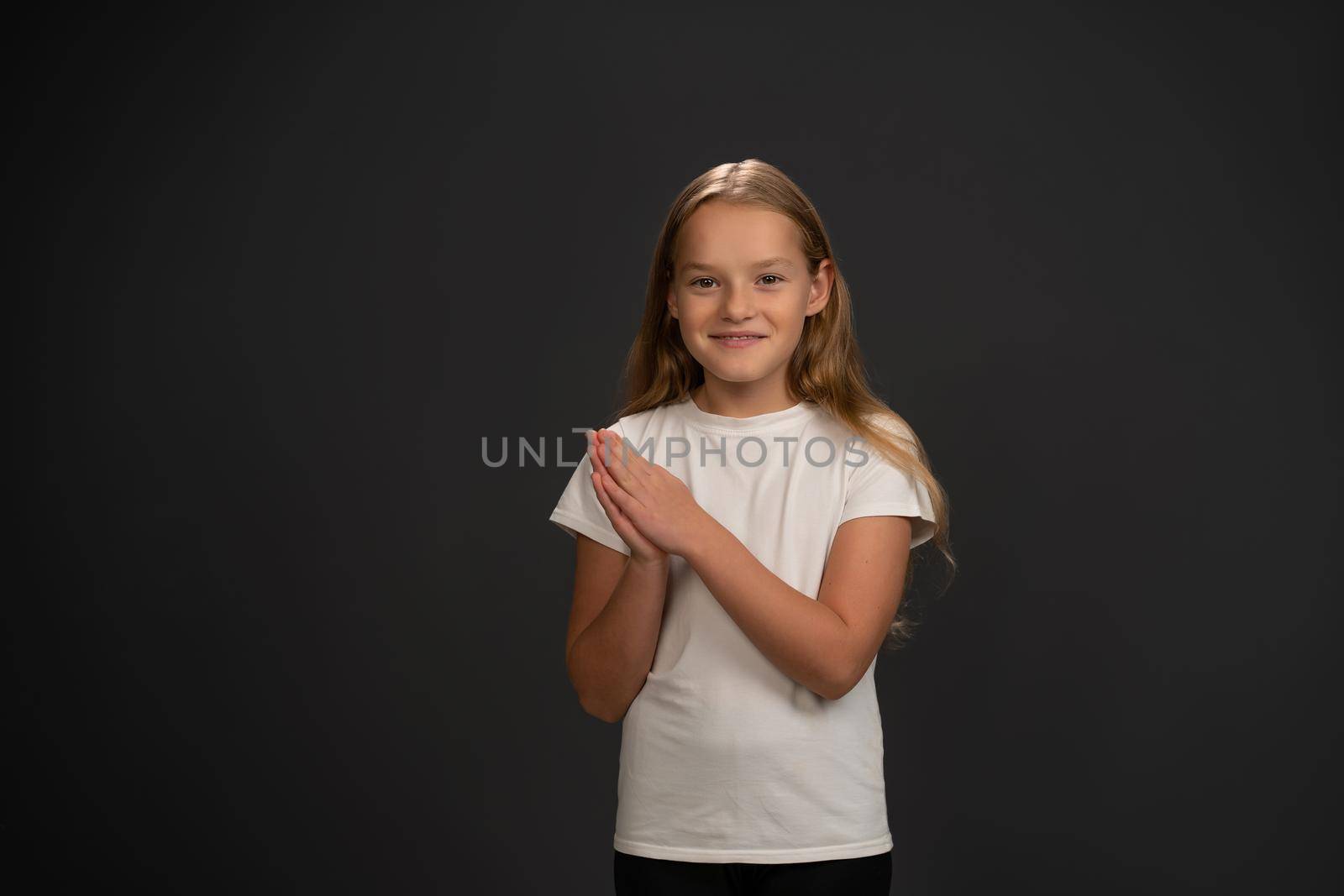 Cheering little girl standing with hands put together happily looking at the camera wearing white t-shirt isolated on dark grey or black background.