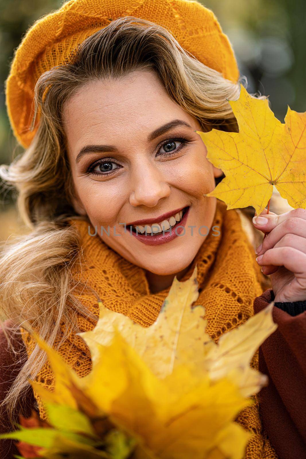 Close up. Young woman in beige knitted beret with autumn leaves in hand and fall yellow garden or park. Beautiful smiling young woman in autumn leaves by LipikStockMedia