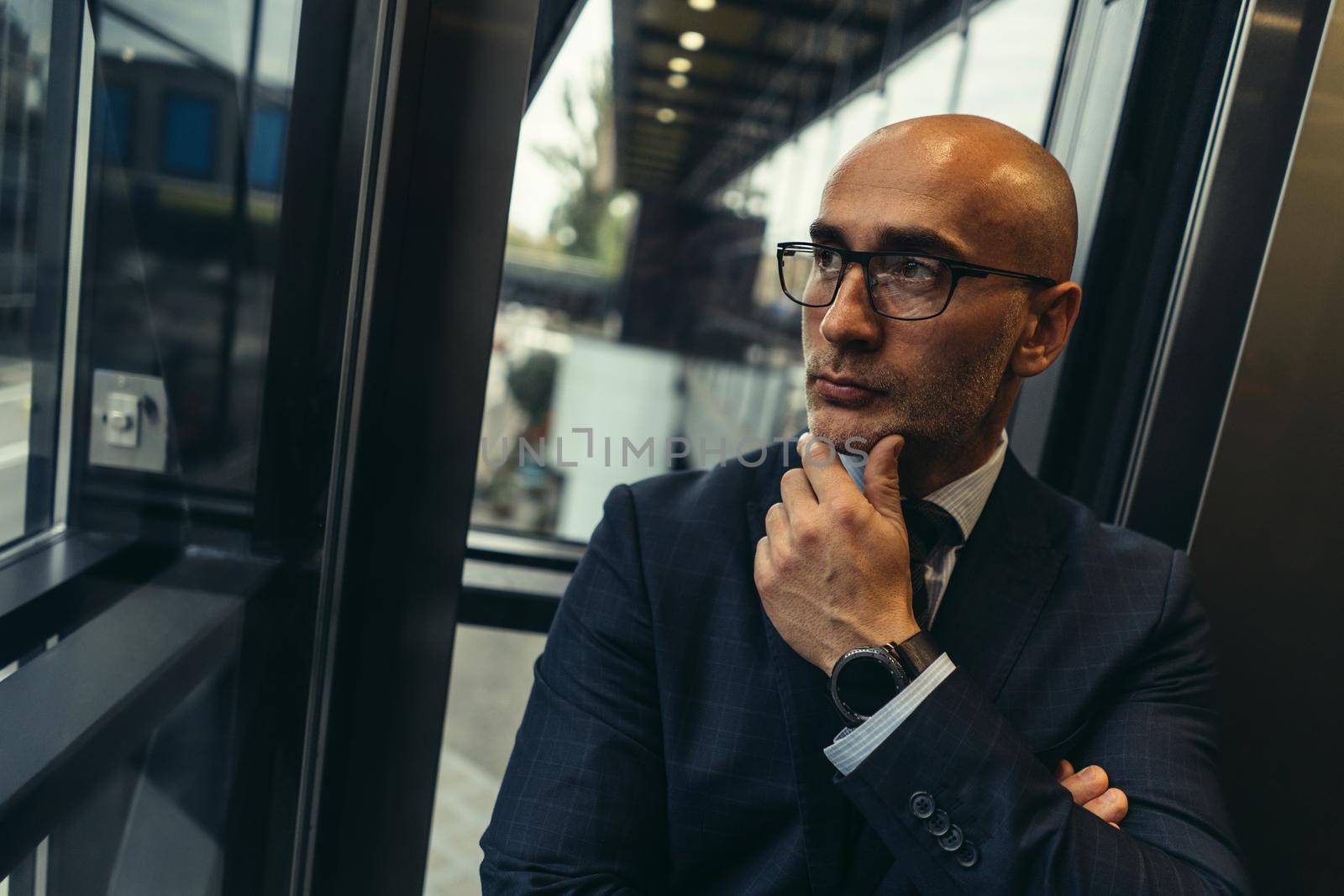 Looking confident with hand raised on chin bald businessman in suit and glasses looking away in the window of modern office with street view. Pensive business man looking outside of the window.
