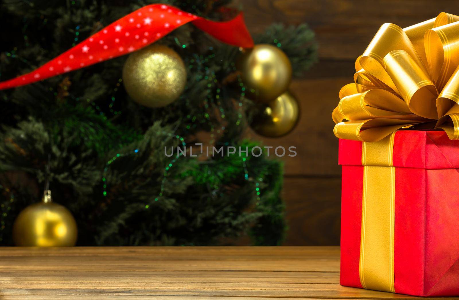 Christmas Gift in a Square Box with a Large Gold Bow Lies on the Table against the Background of a Green Christmas Tree with Gold Balls, Holiday Garlands and a Red Ribbon with Stars. Copy Space. Close-up. High quality photo