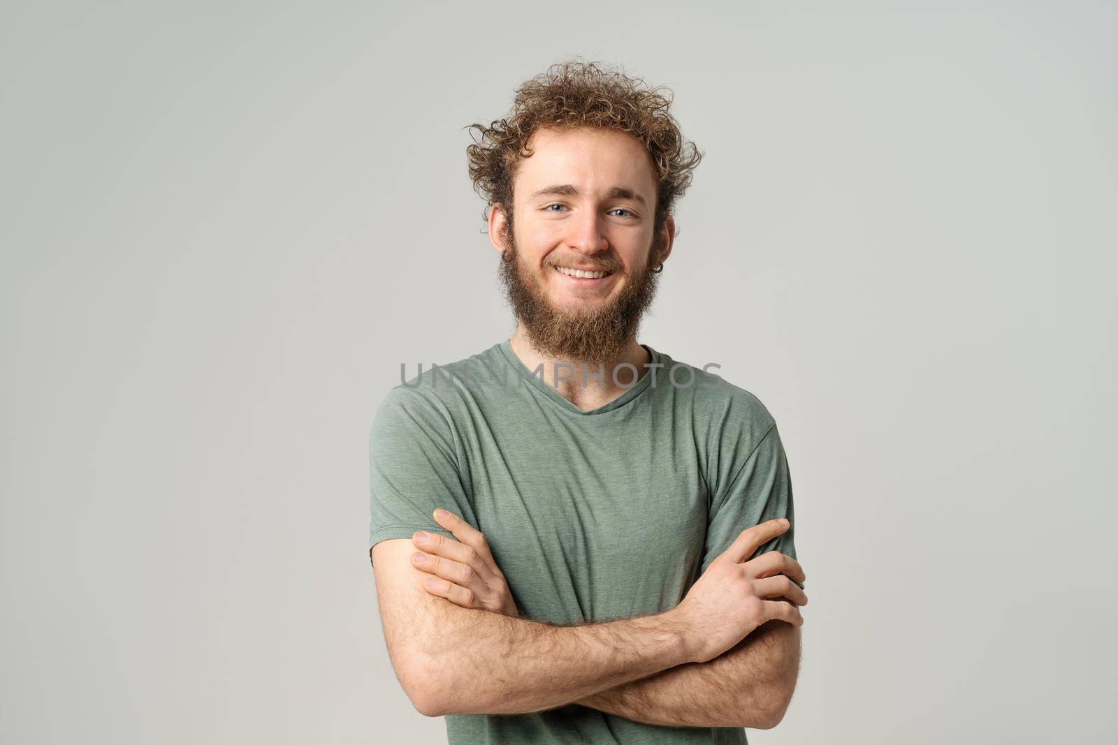 Handsome young man with curly hair in olive t-shirt looking at camera isolated on white background. Portrait of smiling young man with hands folded.