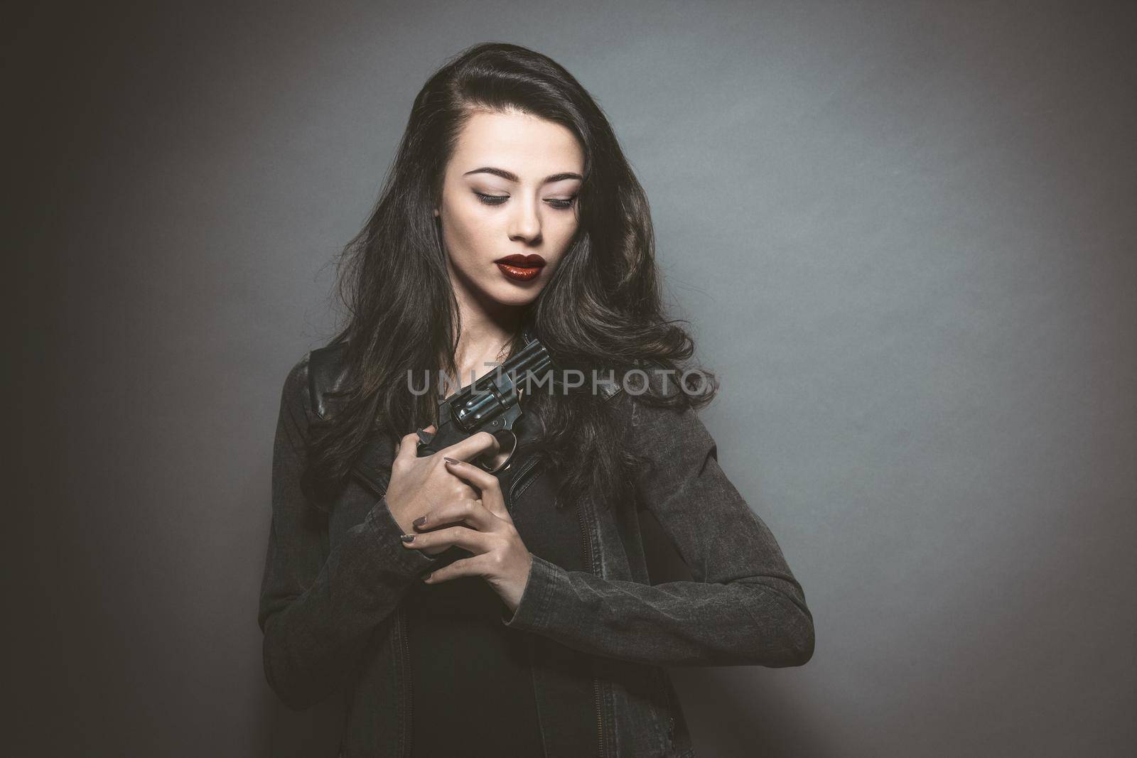 Secret agent beautiful young woman with red lips holding a gun wearing leather jacket. Dangerous young female model with professional make-up. Fashion, beauty, makeup, hairstyle and gun.