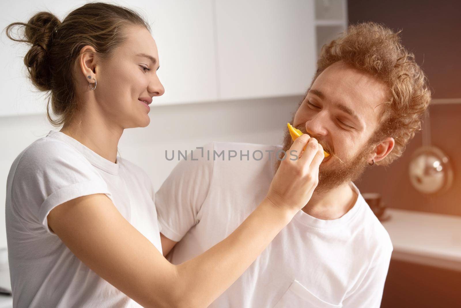 Pretty girl feeds or nursing her boyfriend. Beautiful young couple feeding each other, making fun at modern kitchen and smiling while cooking at home.