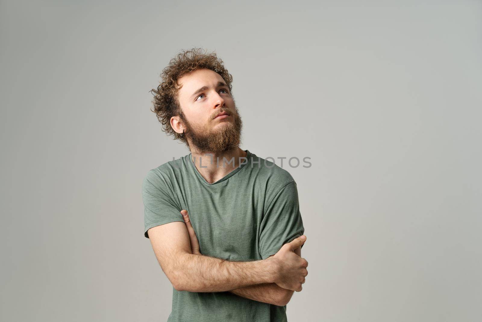 Portrait of pensive deep in thoughts young man with hands folded. Attractive man with curly hair and beard in olive t-shirt looking at camera isolated on white background.