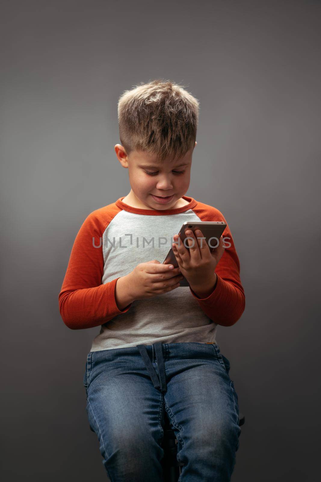 little boy in red shirt looking at the smartphone holding it in his hands and smile isolated on grey background. Human emotions, facial expression concept. Facial expressions, emotions, feelings.
