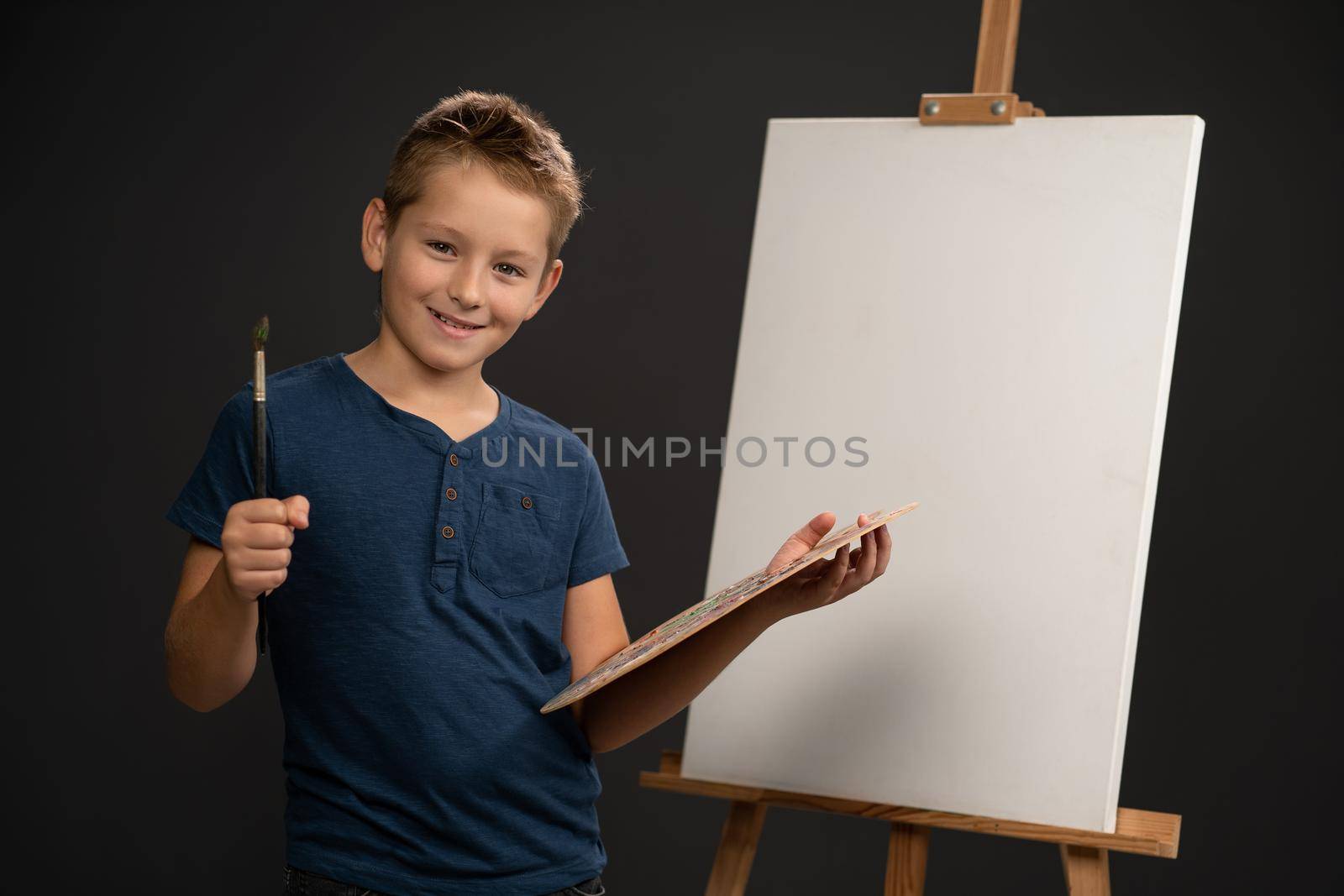Charming boy in blue t-shirt looking at camera holding a palette with paints on the background of an easel with canvas. School of arts concept.