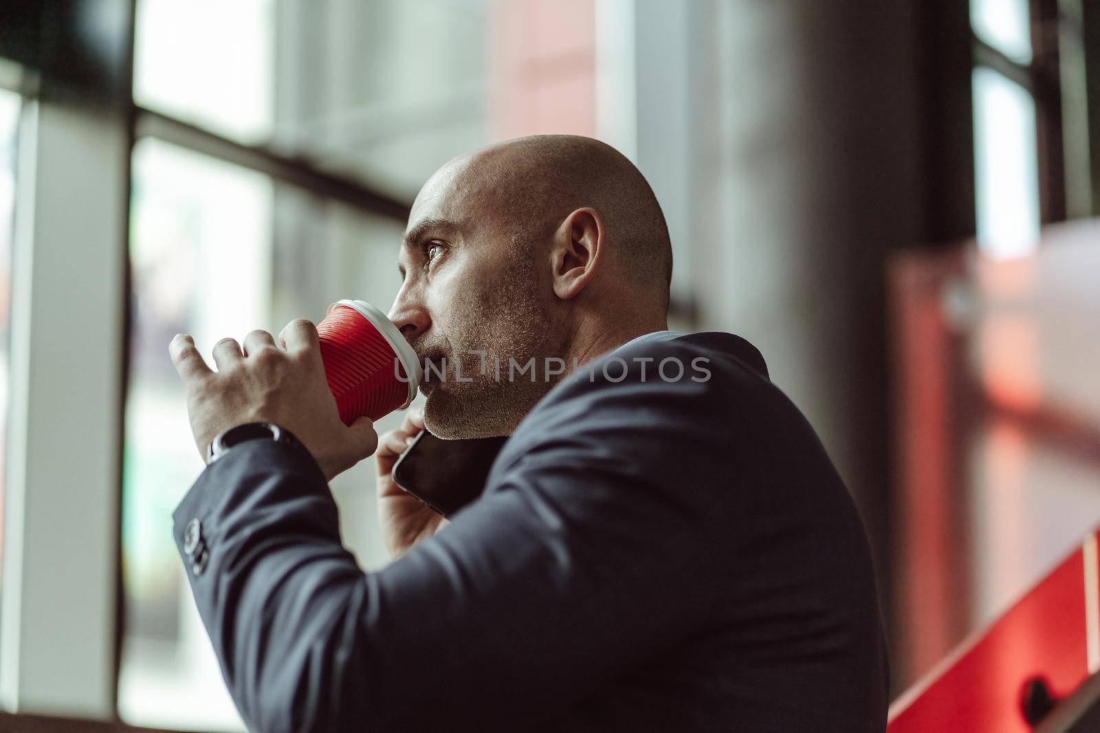 Mature business man in a business suit, talking on the phone while drinking coffee using disposable paper cup standing in a office building. Business concept.