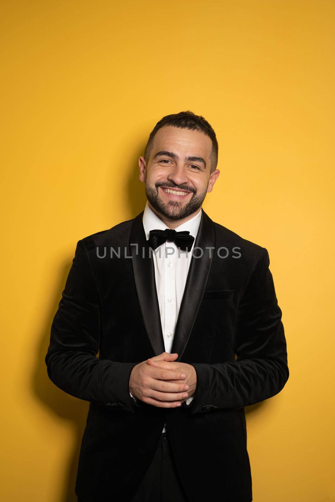 Bearded handsome man in tuxedo with hands folded down smiling on camera, work profession lifestyle. Handsome young smiling caucasian man isolated on yellow background.