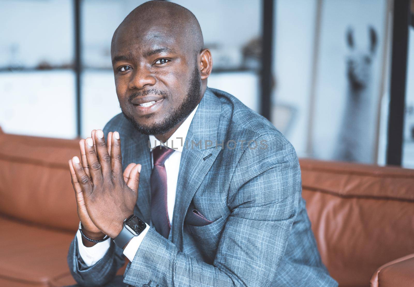 Young Nigerian Guy in a Formal Suit Sits with Folded Hands and Gazes into the Camera. Businessman from Africa is Ready to Provide his Conditions for Cooperation. Close-Up Portrait. Interior Background. High quality photo