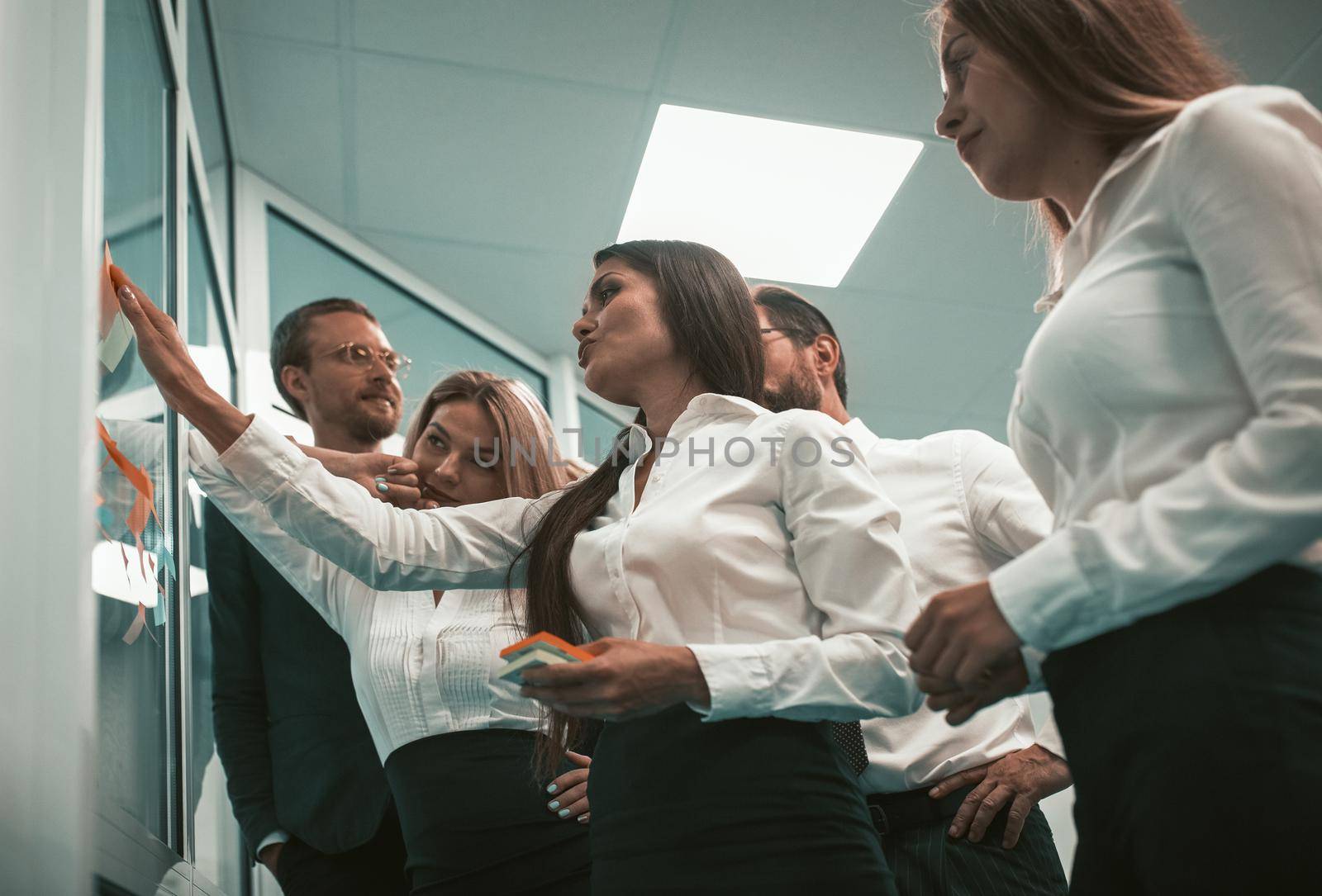 Friendly team conference developing weekly planner, business people brainstorming together looking on colorful stickers on office door window indoors. Low angle view.