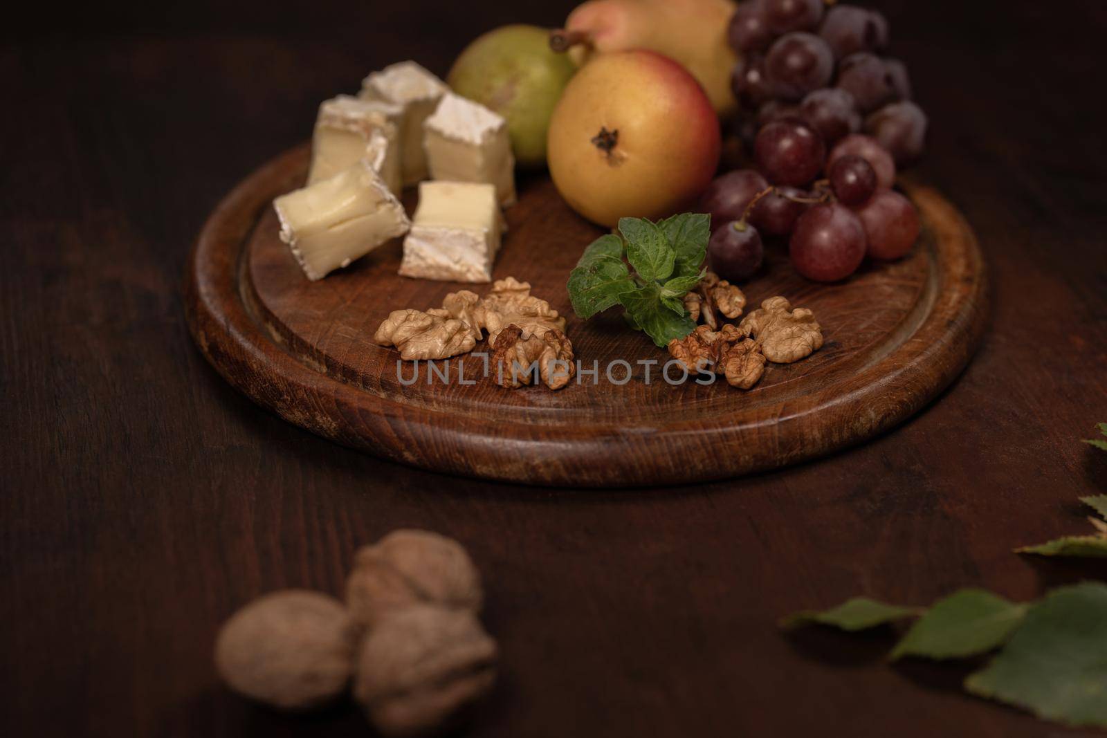 Brie cheese with fruits, grapes, walnuts on a dark wooden plate and wooden board. Wine snacks set: selection of cheese, grapes, pear and three walnuts on a wooden table. Side angle view.