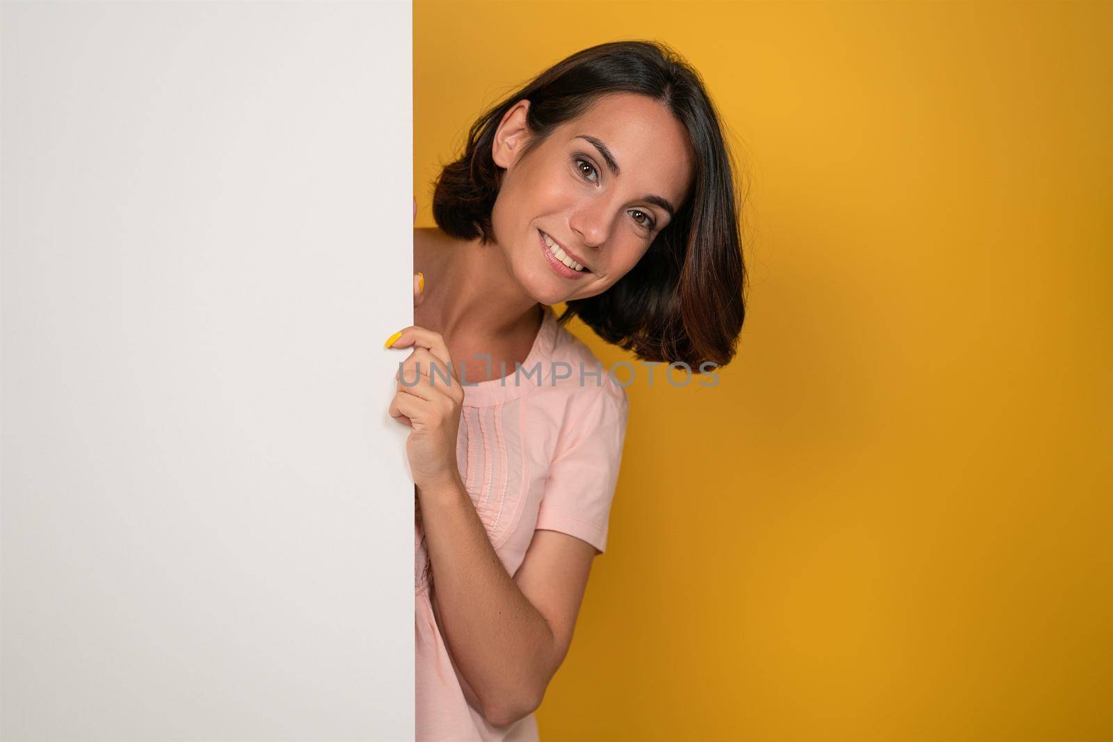 Pretty young woman peeking out of empty banner looks at camera as if watching a family. Smiling young model in a light pink t-shirt looking out of white blank. Isolated on a yellow background.