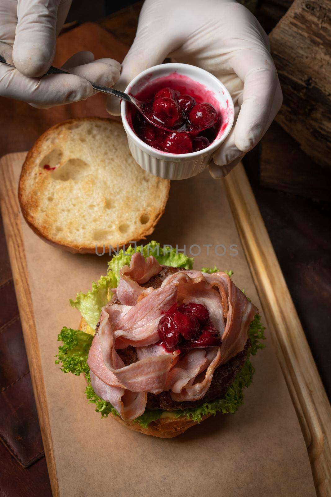 Top view. Chef serving fresh craft beef burger with fried slices of bacon covering with sweet and sour cherry sauce and fresh lettuce before serving it to a client. Street food concept.