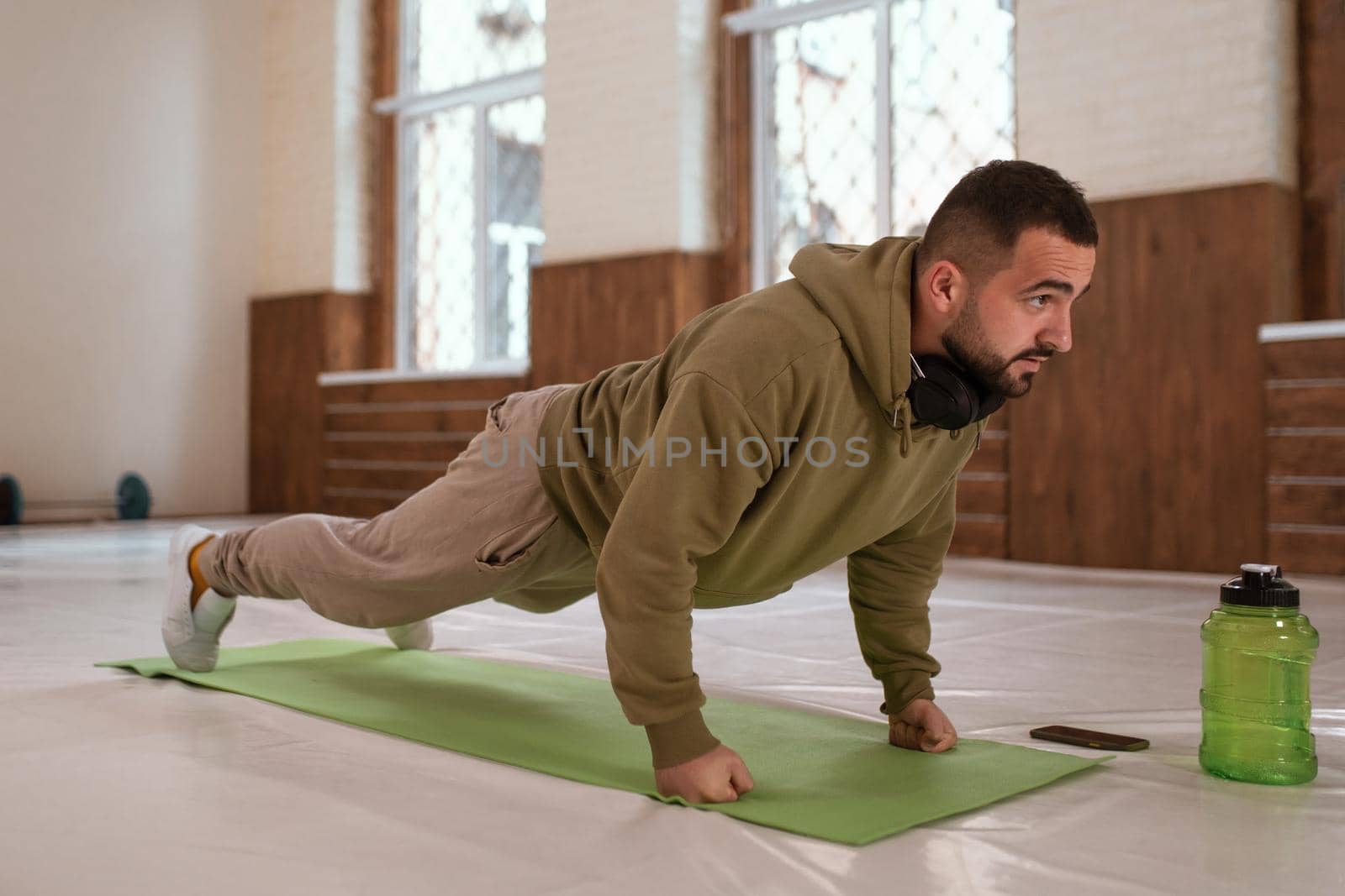 Young handsome sports guy doing push ups exercise in empty gym with green bottle of water next to him all in khaki color clothes. Muscular sportsman doing exercises alone in gym by LipikStockMedia