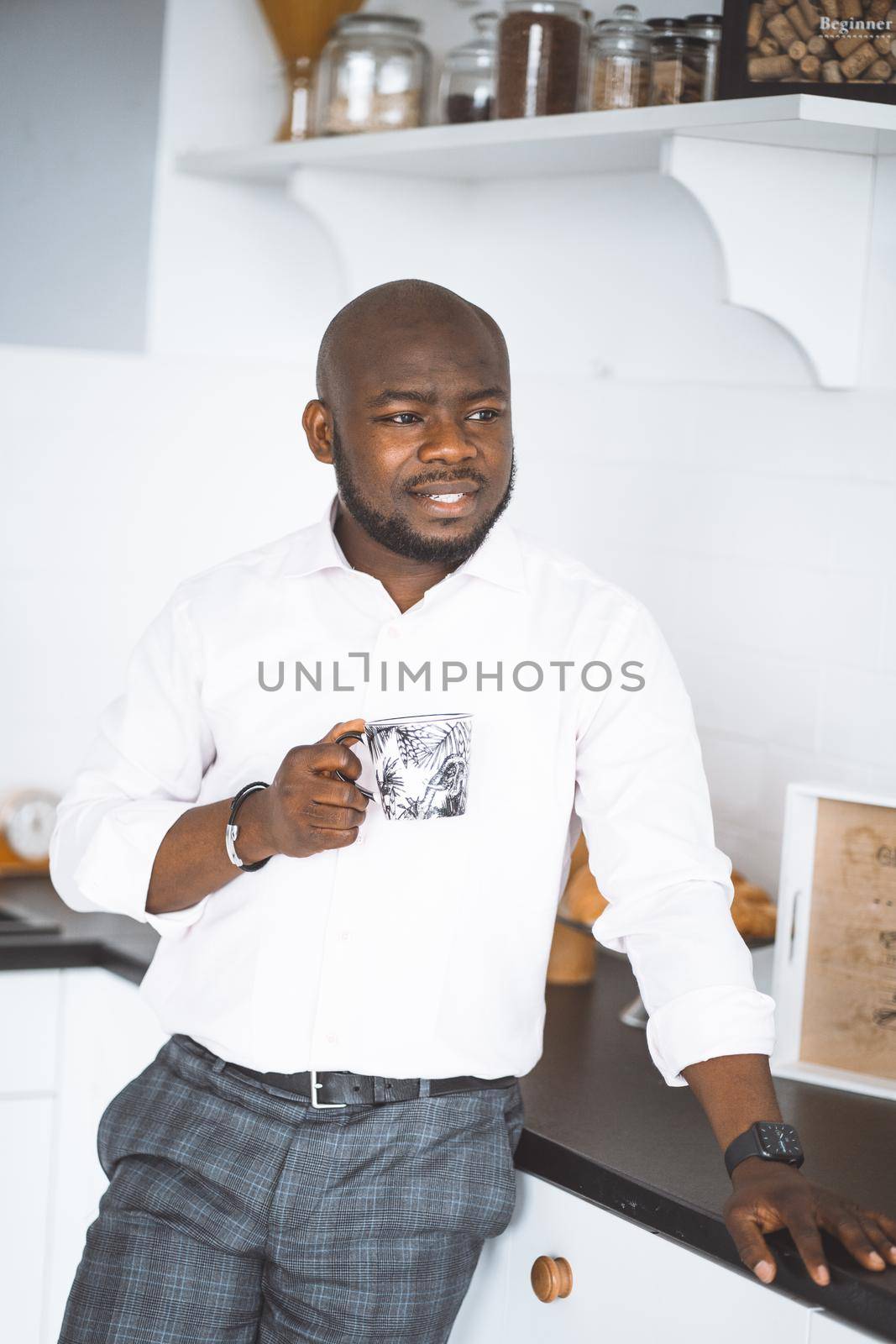 Home Comfort. Afro American Husband With A Cup Stand For In Kitchen After Work by LipikStockMedia