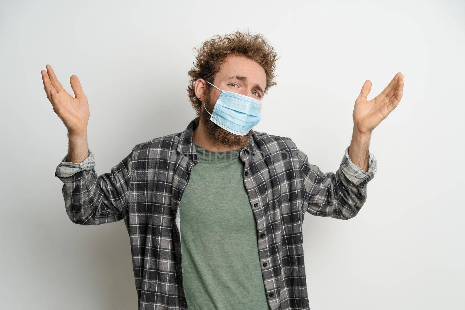 No exit. Young man in protective face mask with curly hair showing I don't know gesture with both hands isolated on white background. Portrait of confused sick young man on white background.