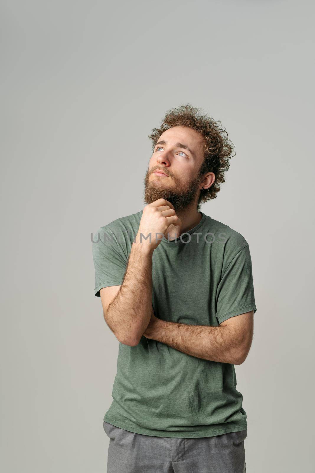 Looking up handsome young man with curly hair in olive t-shirt looking at camera isolated on white background. Portrait of smiling young man with hands folded.