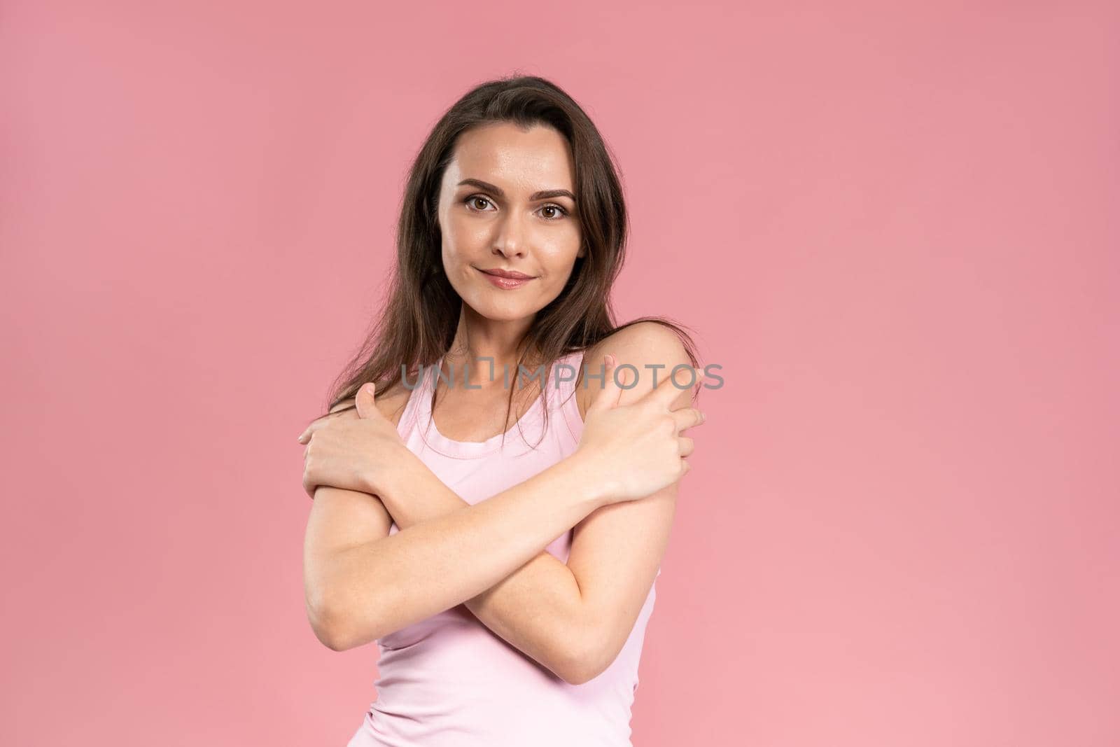 Beautiful young woman with no makeup standing with hands crossed on her chest, attractive brunette girl on pink background. Human emotions, facial expression concept.