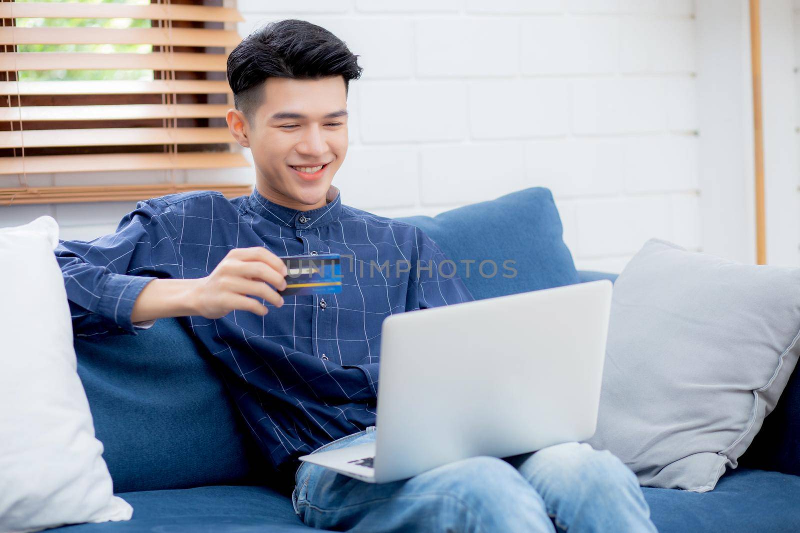 Young asian man sit on sofa using laptop computer shopping online with credit card buying to internet, happy male payment with business on couch, purchase and payment, business concept. by nnudoo