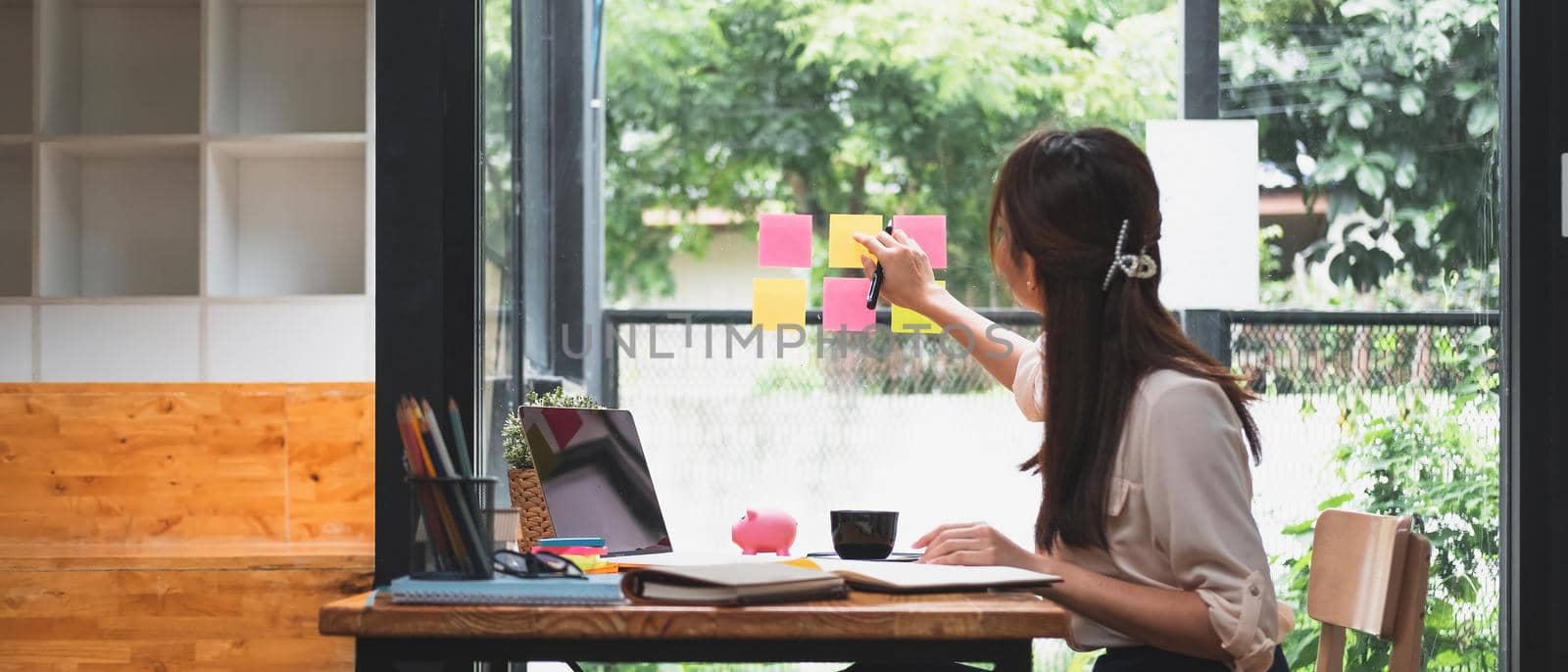 Business attractive young asian woman write notes on sticky note paper to arrange meeting with coworker sit in background take pictures through the glass, start up business and financial concept.