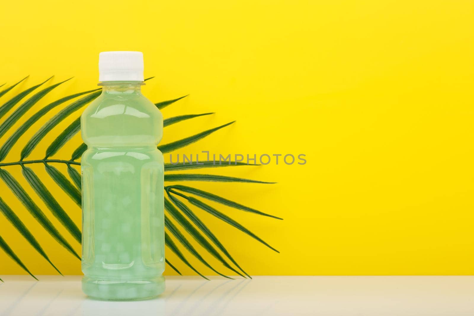 Detox drink or juice in transparent bottle with small pieces of fruits on white table against yellow background with palm leaf and copy space. Concept of healthy eating, dieting, weight loss and detox 