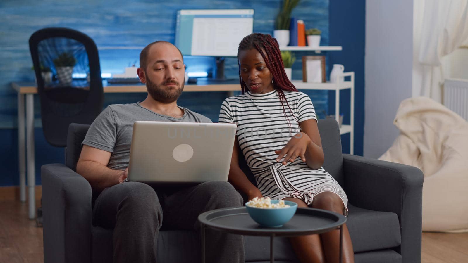 Young interracial couple looking at modern laptop sitting together in living room. African american woman and caucasian man using technology at home for entertainment and bonding