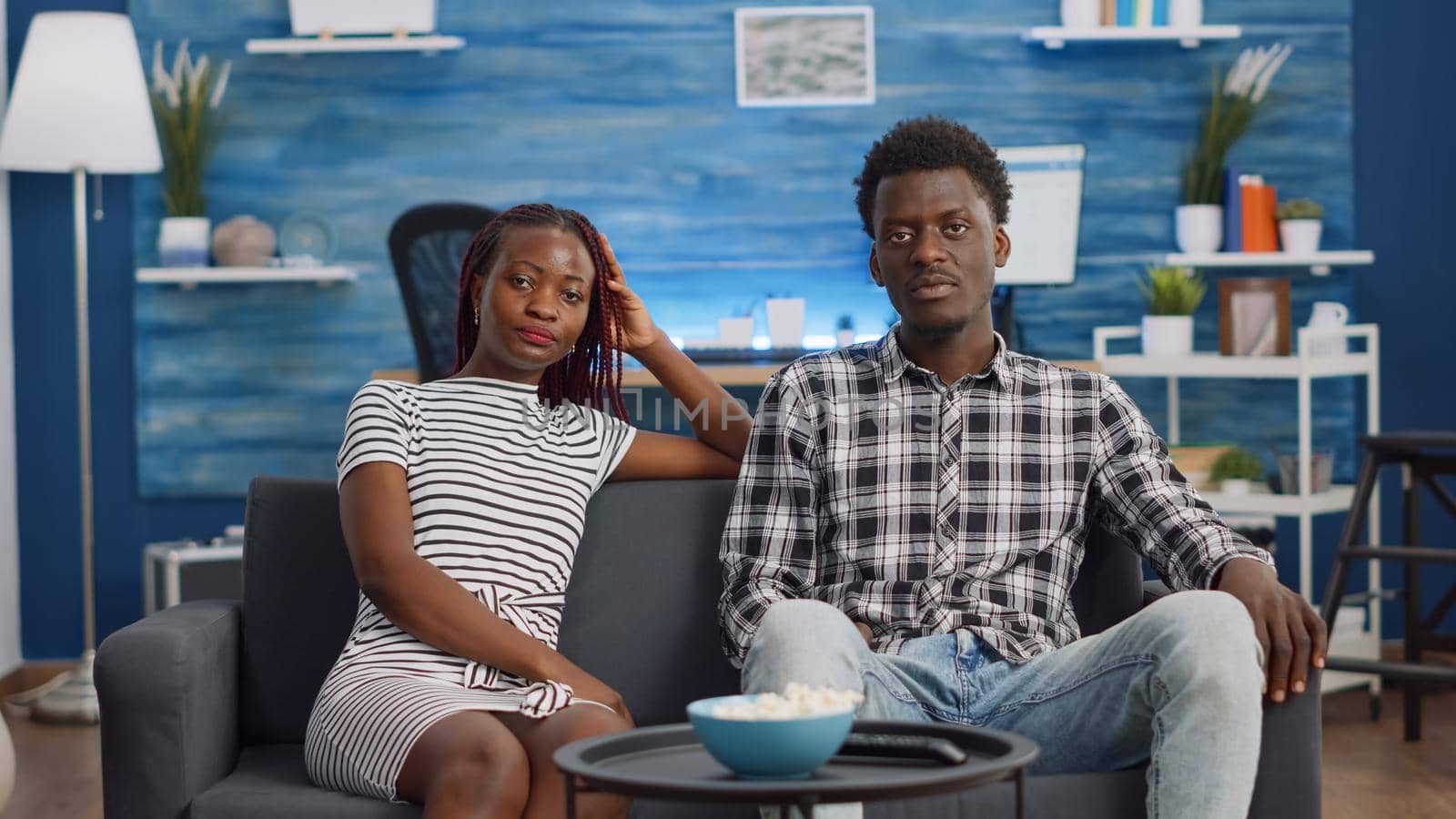 African american couple watching scary movie on TV, sitting on couch. Black people looking at camera, young woman covering eyes being afraid while man laughing. Lovers enjoying time together