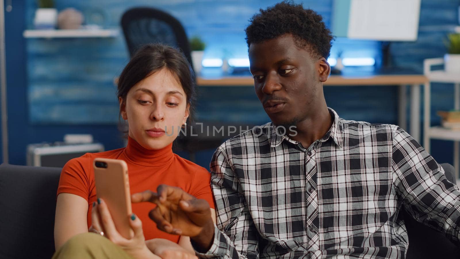 Modern interracial couple using smartphone in living room. Young multi ethnic people looking at digital device and relaxing on couch at home. Cheerful husband and wife sitting together