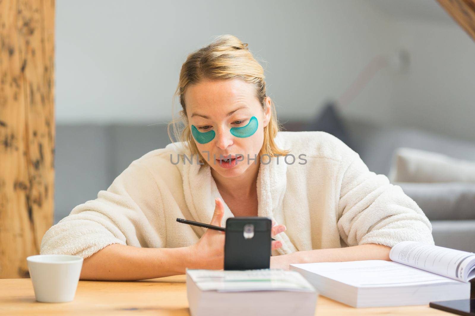 Young businesswoman wearing cosy warm bathrobe having remote cell phone conference call meeting from home living room in winter Christmas time. Remote work from home concept
