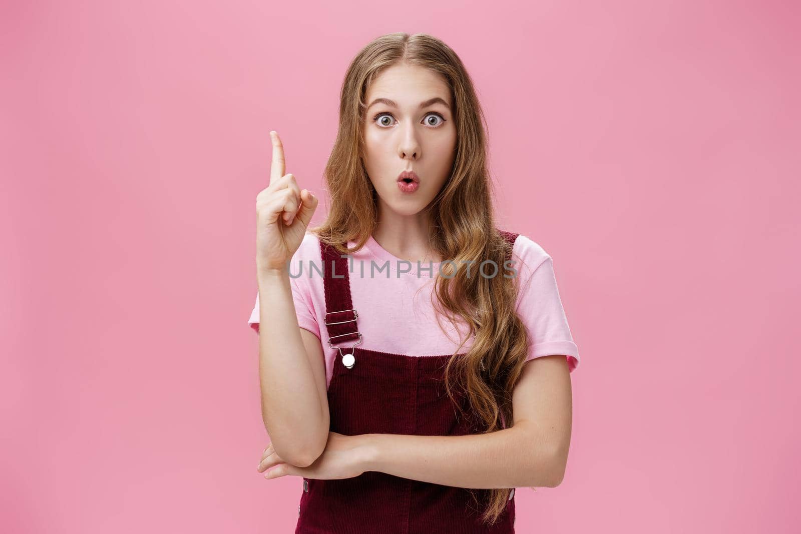 Young smart and creative female student sharing with team awesome idea coming in mind raising index finger in eureka gesture looking excited at camera folding lips saying suggestion over pink wall. Body language, body-positive and gestures concept