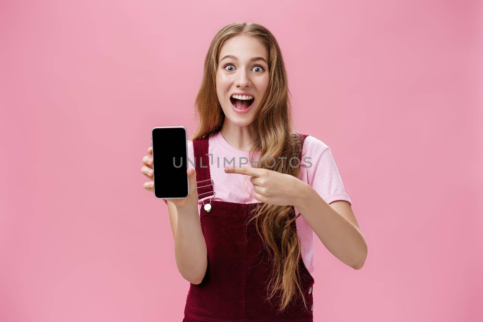 Excited and thrilled young teenage girl in overalls and t-shirt smiling amazed full of happiness pointing at smartphone screen staring enthusiastic at camera describing awesome cellphone features. Technology concept