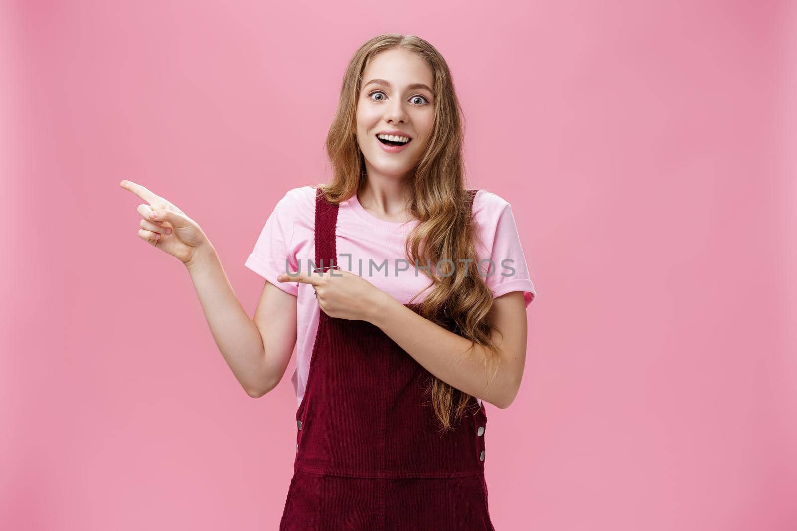 Lifestyle. Woman looking with impressed and amazed look showing cool think on left side of copy space smiling fascinated looking with admiration at camera standing delighted and surprised over pink background.