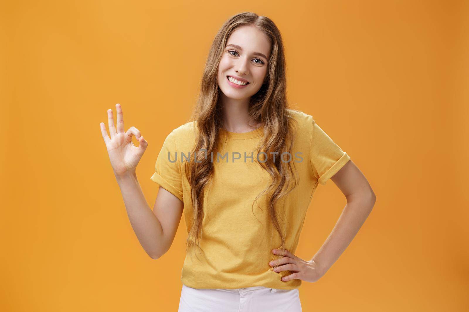 Girl showing positive answer with body language. Charming friendly-looking young female student with natural wavy hairstyle holding hand on hip making ok gesture and smiling over orange wall by Benzoix