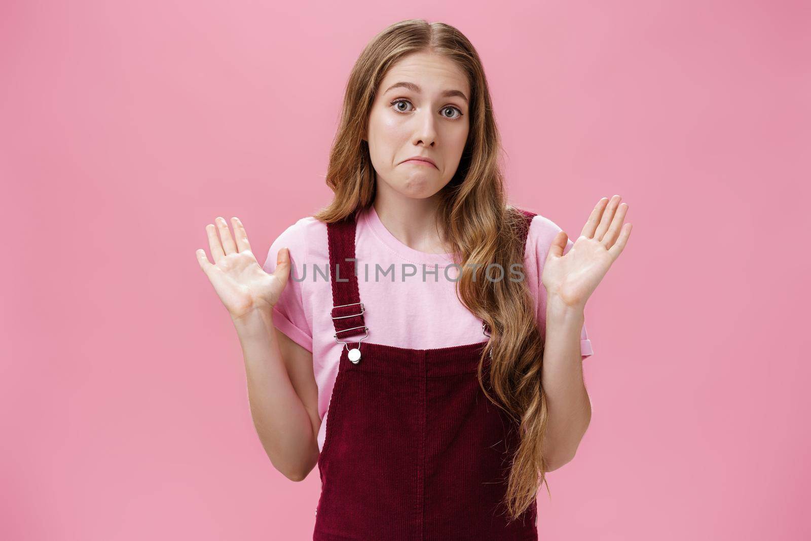 I am not involved. Portrait of unaware and clueless innocent cute woman raising palms in surrender pulling lips down as if being confused and uncertain what happening having no idea over pink wall by Benzoix