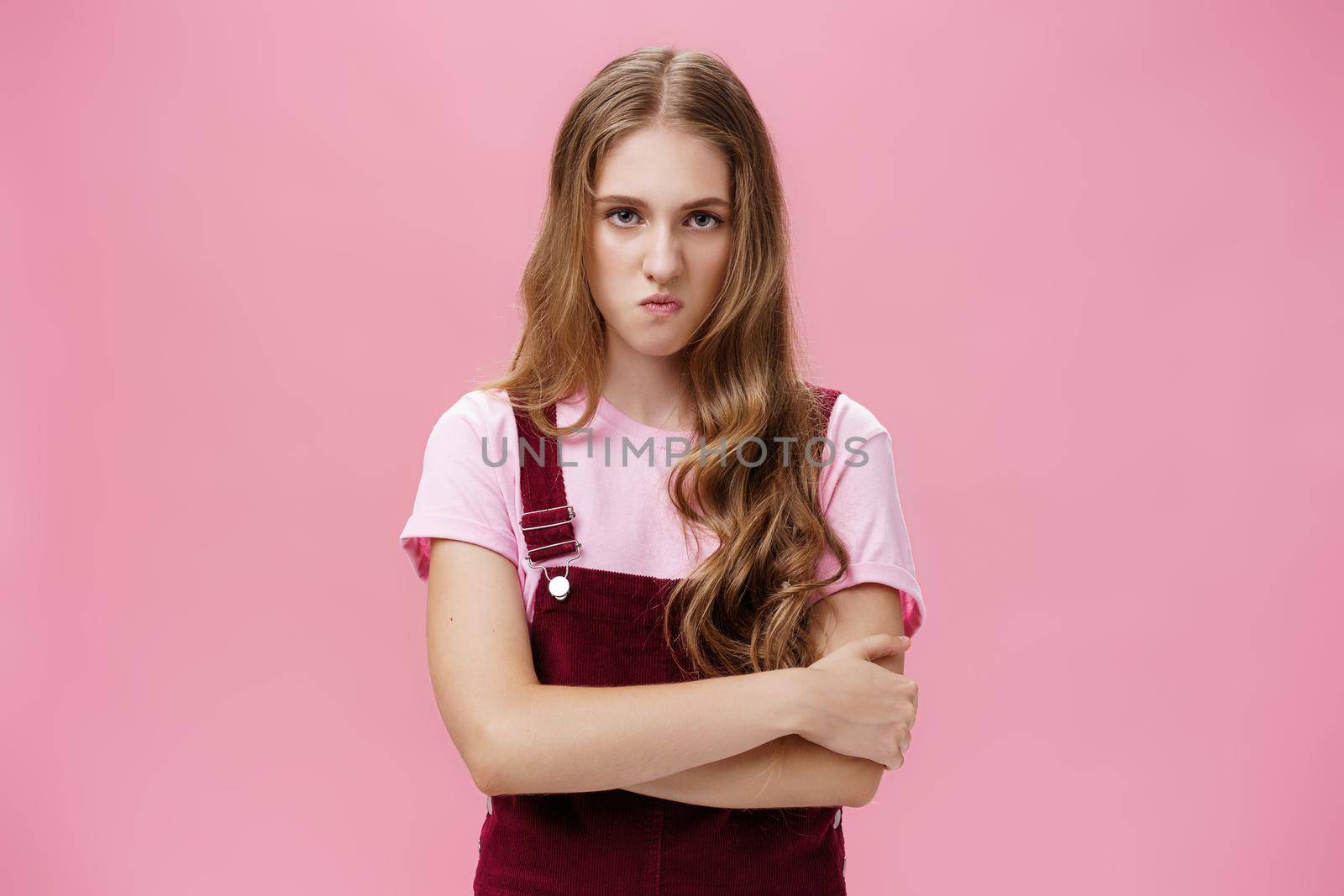 I mad on world. Unhappy hateful young arrogant girl in overalls with wavy natural hairstyle crossing arms against chest in defensive pose lifting upper lip in scorn and disdain, looking with anger by Benzoix