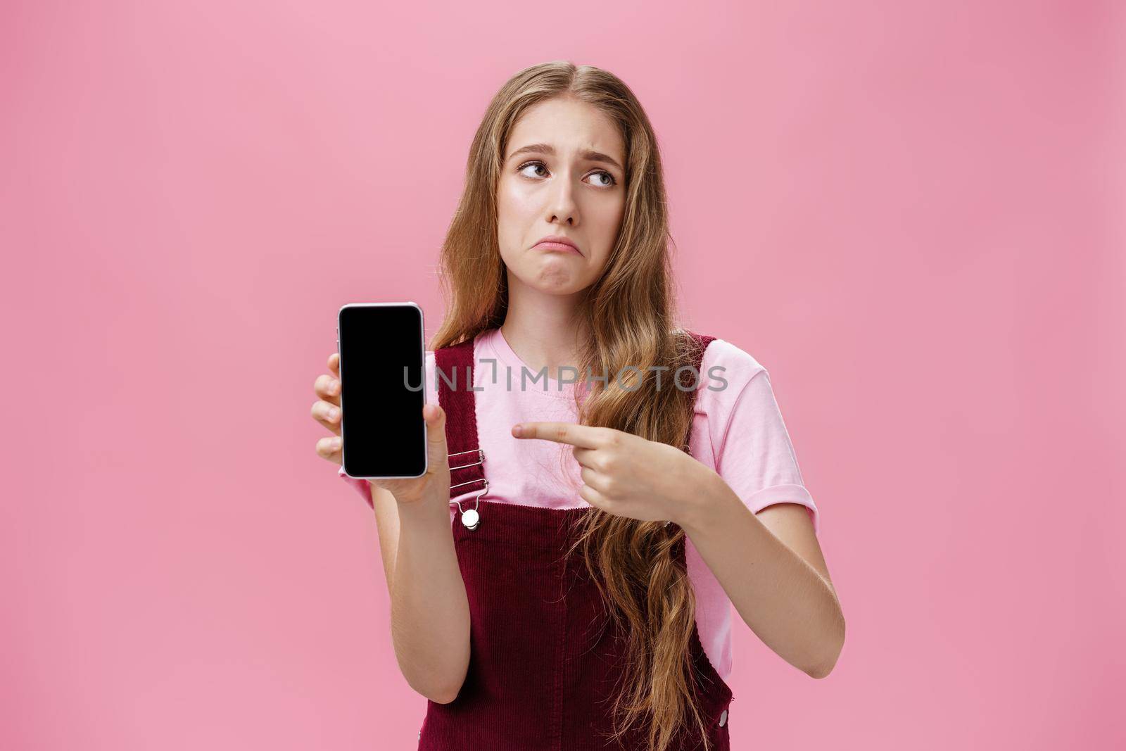 Indoor shot of gloomy displeased and disappointed cute young girl holding smartphone pointing at cellphone screen making upset grimace as if being bothered and dissatisfied against pink wall by Benzoix